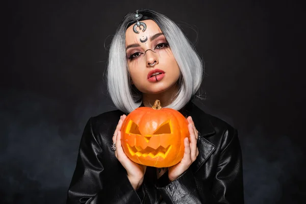 Young ash blonde woman with witch makeup holding jack o lantern on black background — Stock Photo