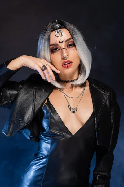 Sexy mujer en traje de bruja y maquillaje sosteniendo la mano cerca de la cara en negro y azul de fondo - foto de stock