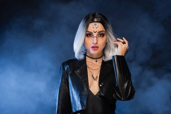 Sexy ash blonde woman in leather jacket and halloween makeup looking at camera on dark background with blue fog — Stock Photo