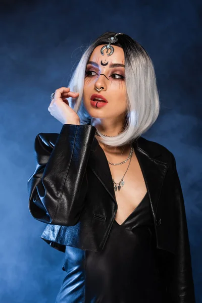 Sexy ash blonde woman in witch makeup looking away on dark background with blue smoke — Stock Photo