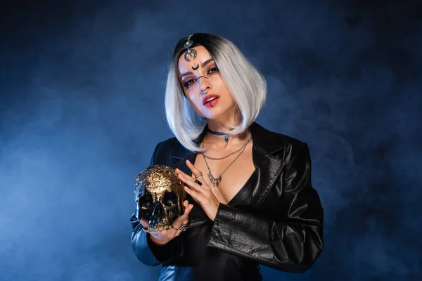 Sexy mujer en halloween traje celebración de cráneo de oro sobre fondo oscuro con niebla azul - foto de stock