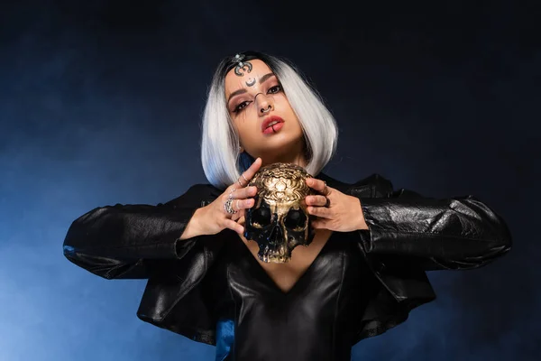 Woman in leather jacket and halloween makeup posing with skull on dark and blue background — Stock Photo
