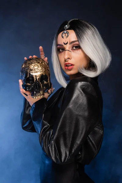 Sexy Hexe Stil Frau mit gruseligen goldenen Totenkopf Blick in die Kamera in der Nähe blauer Nebel auf dunklem Hintergrund — Stockfoto