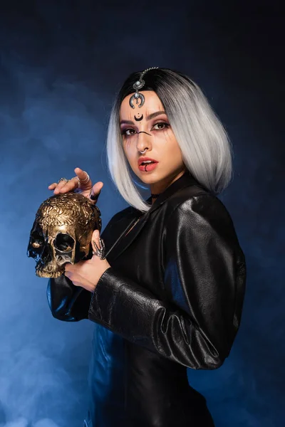 Ash blonde woman in halloween makeup and leather jacket holding golden skull on black background with blue smoke — Stock Photo