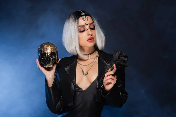 Ash blonde woman in halloween costume holding black rose and golden skull on dark background with blue fog — Stock Photo