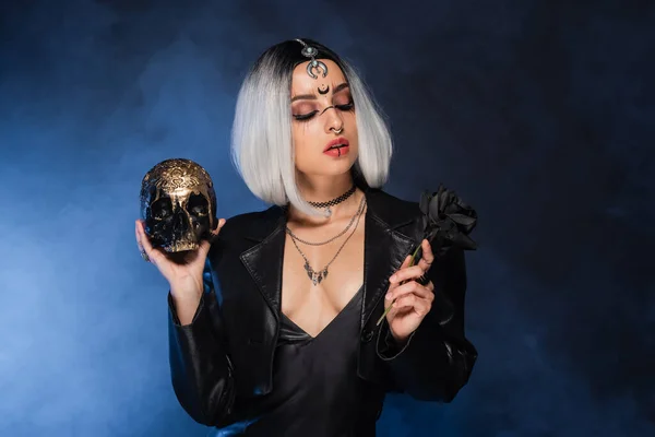 Aschblonde Frau im Halloween-Kostüm mit schwarzer Rose und goldenem Totenkopf auf dunklem Hintergrund mit blauem Nebel — Stockfoto