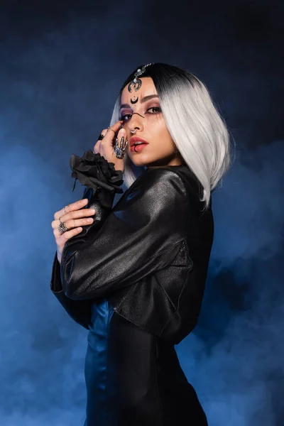 Seductive woman in witch costume holding black rose and looking at camera on dark background with blue smoke — Stock Photo