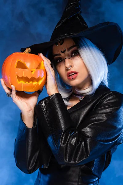 Mulher em bruxa halloween traje segurando esculpida abóbora no azul embaçado fundo — Fotografia de Stock