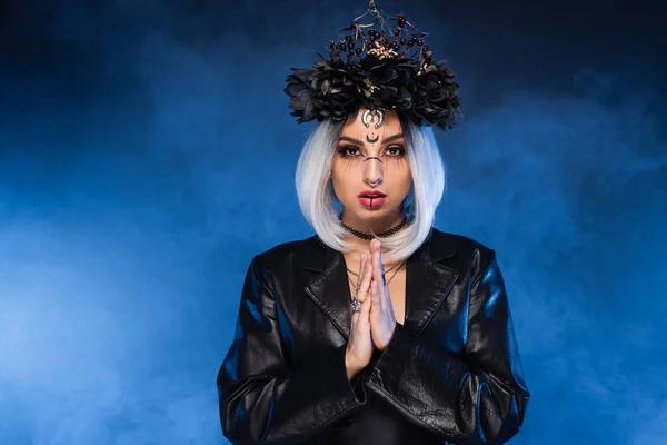 Femme de style sorcière en couronne noire et veste en cuir debout avec les mains priantes sur fond sombre avec brouillard bleu — Photo de stock