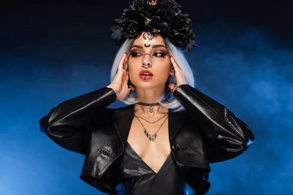 Sexy woman in black wreath and witch makeup touching ash blonde hair on dark background with blue smoke — Stock Photo