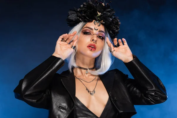 Sexy mujer en bruja halloween traje y corona tocando ceniza pelo rubio sobre fondo azul oscuro - foto de stock