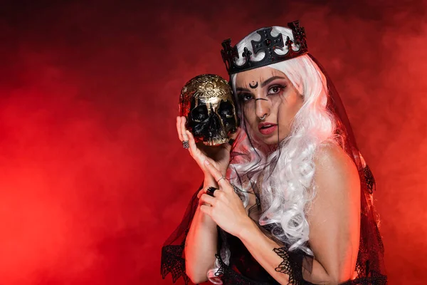 Blonde Frau im Hexenstil mit schwarzer Krone und Schleier blickt in die Kamera in der Nähe des goldenen Totenkopfes auf rotem rauchigen Hintergrund — Stockfoto