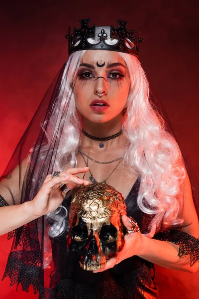 Blonde woman in witch makeup and crown holding golden skull and looking at camera on red — Stock Photo