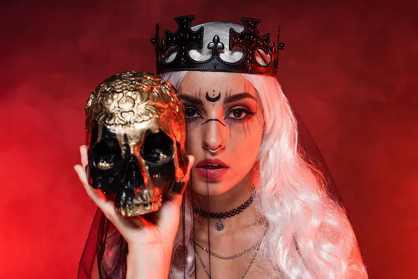 Mulher loira com maquiagem bruxa vestindo coroa preta e véu enquanto olha para a câmera perto do crânio dourado no fundo vermelho — Fotografia de Stock