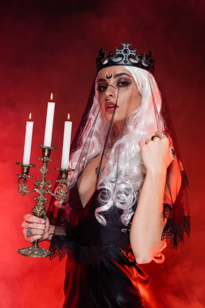 Sexy femme blonde en costume de sorcière debout avec triple chandelier sur fond sombre avec brouillard rouge — Photo de stock