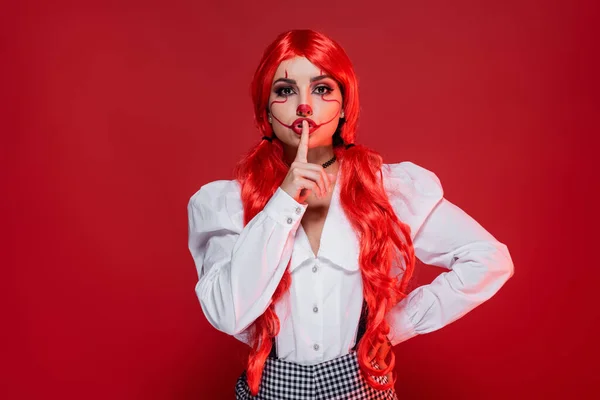 Mujer con maquillaje estilo payaso mostrando signo de silencio mientras está de pie con la mano en la cadera aislado en rojo - foto de stock