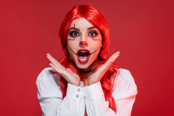 Mujer pelirroja asombrada con el maquillaje de Halloween cogido de la mano cerca de la cara aislado en rojo - foto de stock