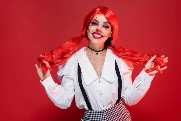 Cheerful woman with clown halloween makeup holding bright ponytails isolated on red — Stock Photo