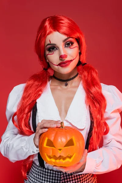 Mulher sorridente com rabo de cavalo brilhante e maquiagem halloween segurando jack o lanterna isolada no vermelho — Fotografia de Stock