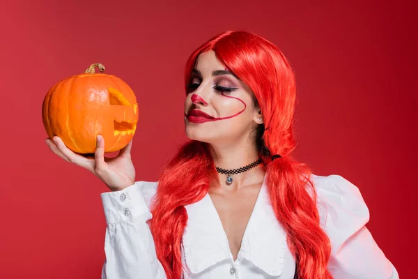 Redhead woman with halloween makeup looking at jack o lantern isolated on red — Stock Photo