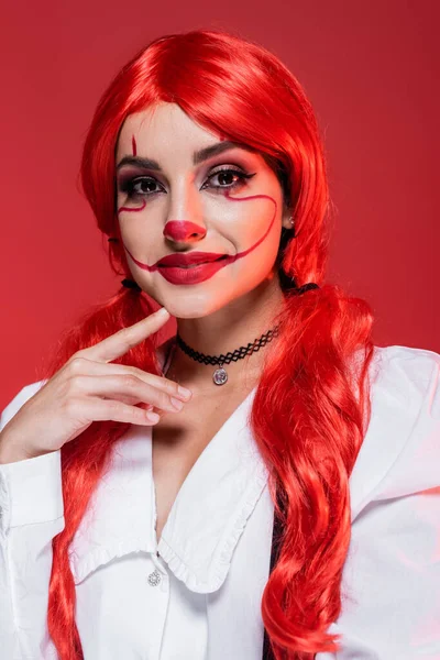 Retrato de mulher sorridente com maquiagem de palhaço e cabelo brilhante tocando queixo e olhando para a câmera isolada no vermelho — Fotografia de Stock