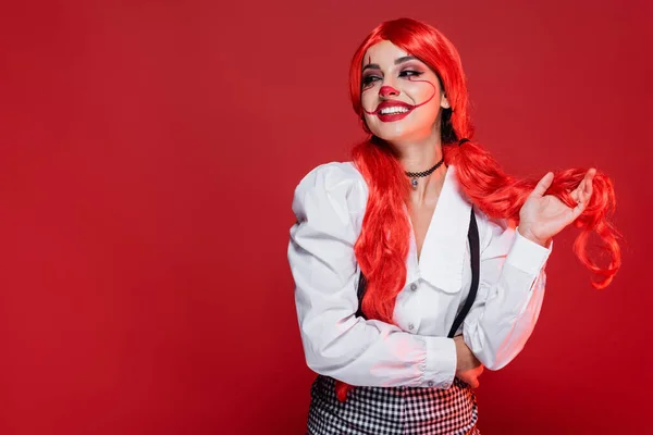 Alegre pelirroja en maquillaje de payaso y blusa blanca mirando hacia otro lado aislado en rojo - foto de stock