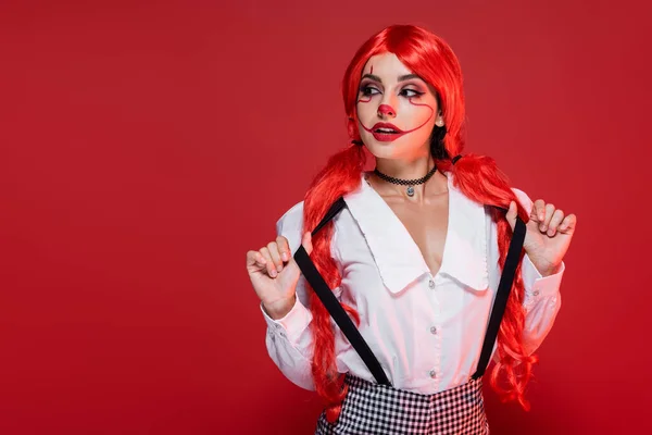 Junge rothaarige Frau in Halloween-Make-up berührt Hosenträger und sieht isoliert auf rot — Stockfoto