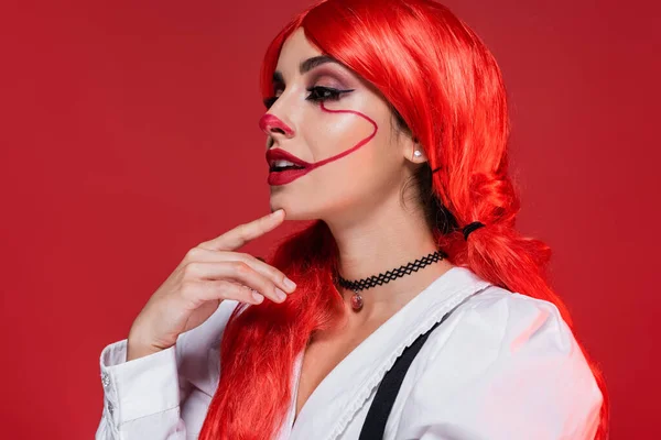 Mujer reflexiva y sonriente con pelo brillante y maquillaje payaso mirando hacia otro lado aislado en rojo - foto de stock