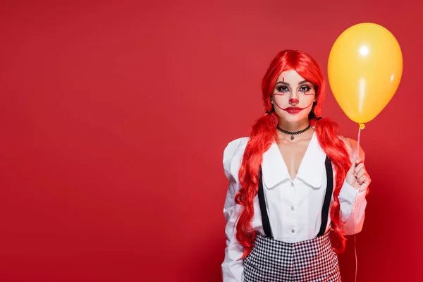 Jovem ruiva em blusa branca e maquiagem de palhaço segurando balão amarelo isolado no vermelho — Fotografia de Stock