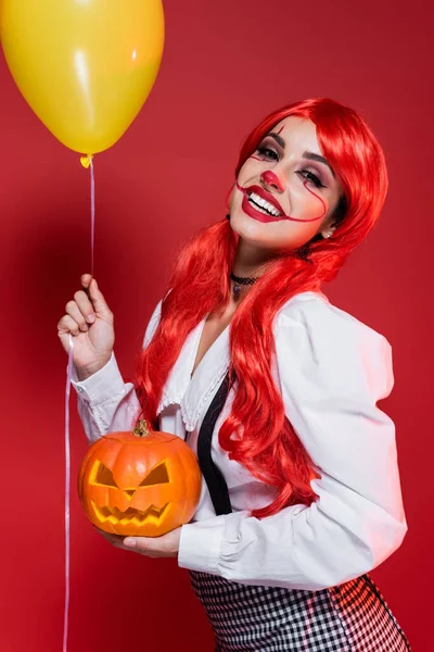 Mujer alegre con el pelo brillante y el maquillaje de Halloween sosteniendo globo amarillo y calabaza tallada aislada en rojo - foto de stock