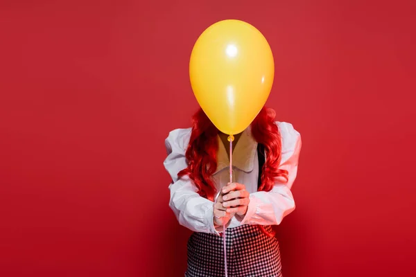 Young woman with colored hair obscuring face with yellow balloon on halloween party isolated on red — Stock Photo