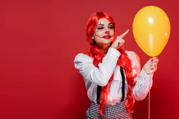 Happy woman with bright hair and clown makeup pointing at yellow balloon isolated on red — Stock Photo