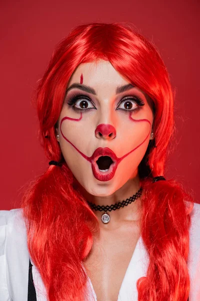 Pelirroja mujer asombrada con el maquillaje de Halloween mirando a la cámara aislada en rojo - foto de stock