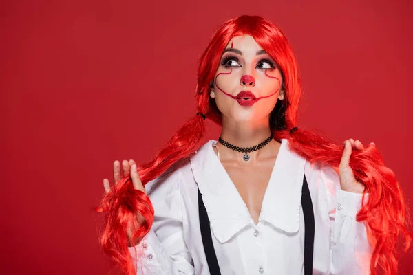 Tricky woman with clown makeup holding bright ponytails and looking up isolated on red — Stock Photo