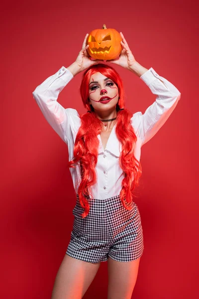Молодая рыжая женщина в Хэллоуин макияж с резной тыквой над головой изолированы на красный — стоковое фото