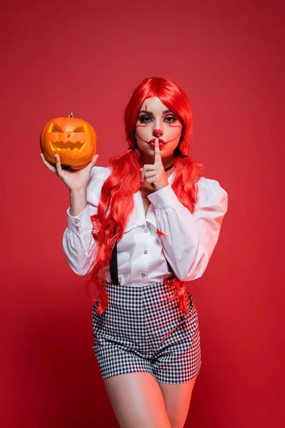 Mulher com maquiagem de palhaço e cabelo colorido mostrando sinal de silêncio enquanto segurando jack o lanterna isolada no vermelho — Fotografia de Stock