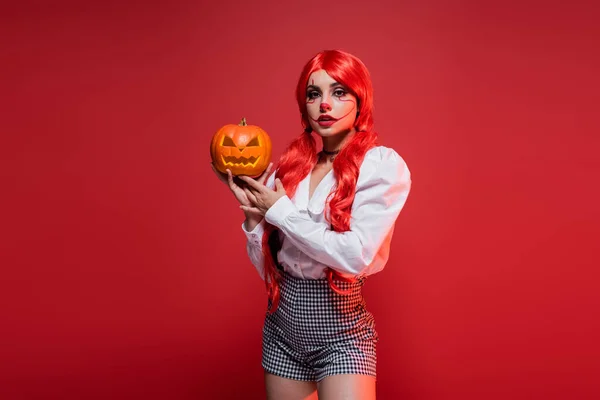 Femme avec des queues de cheval brillantes et maquillage de clown tenant la citrouille sculptée isolée sur rouge — Photo de stock