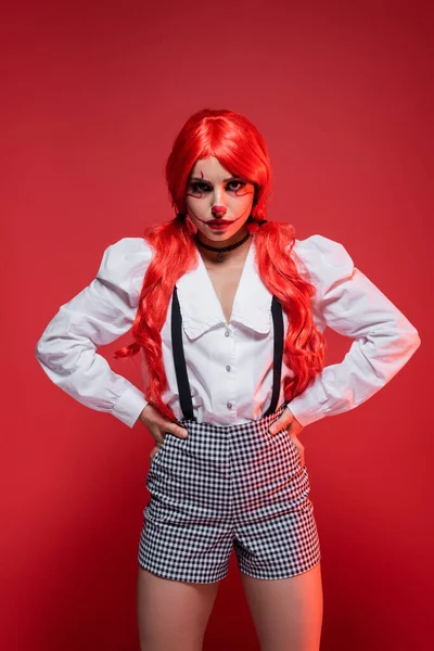 Femme en colère avec des cheveux longs brillants et maquillage de clown debout avec les mains sur les hanches isolées sur rouge — Photo de stock