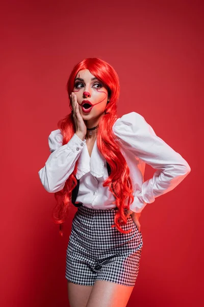 Mulher ruiva espantada em halloween traje tocando rosto e de pé com a mão no quadril isolado no vermelho — Fotografia de Stock