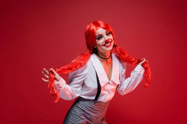 Sorrindo e flertando mulher segurando rabos de cavalo brilhantes e olhando para a câmera isolada no vermelho — Fotografia de Stock