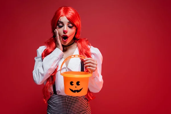 Amazed woman with bright hair and clown makeup looking in halloween bucket isolated on red — Stock Photo