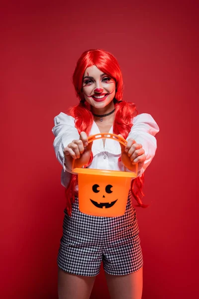 Alegre mujer en payaso maquillaje y brillante peluca mostrando halloween cubo aislado en rojo - foto de stock