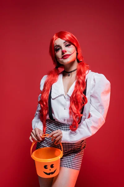 Mulher com cabelo colorido e maquiagem de palhaço posando com balde halloween isolado no vermelho — Fotografia de Stock