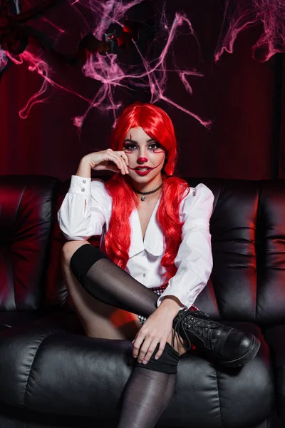 Sexy woman in black knee socks and halloween makeup looking at camera on leather couch and black background with spiderweb — Stock Photo