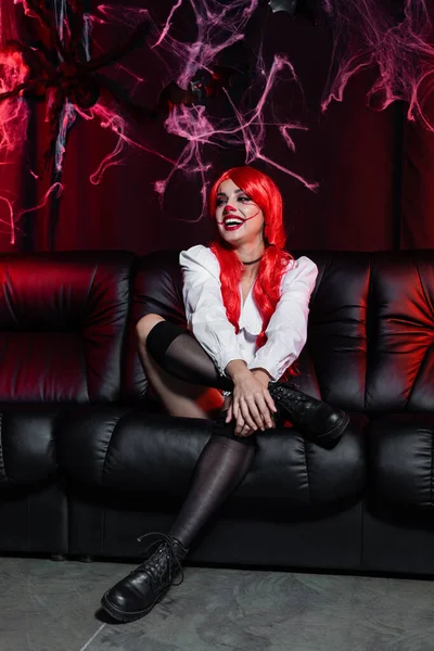 Mulher alegre e sexy na maquiagem halloween sorrindo no sofá de couro perto teia de aranha no fundo preto — Fotografia de Stock