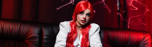 Рыжая женщина с хвостиками и хэллоуин макияж на черном диване и темный фон с паутиной, баннер — стоковое фото