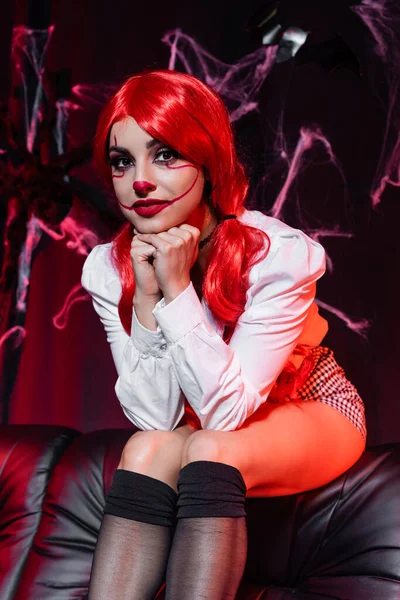 Sexy woman in clown costume looking at camera while sitting on dark background with spiderweb — Stock Photo