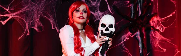 Redhead woman in clown makeup holding skull mask near spiderweb on dark background, banner — Stock Photo