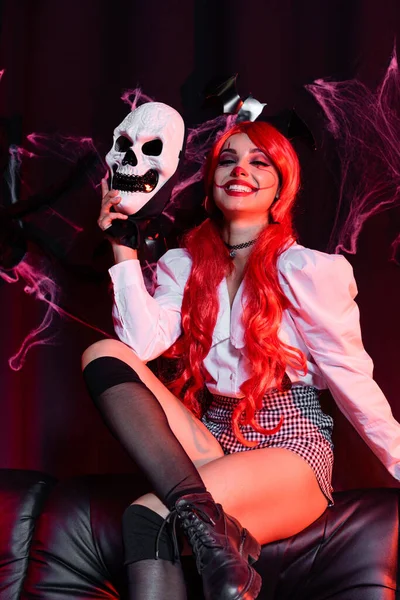 Cheerful redhead woman in clown makeup holding skull mask on black background with spiderweb — Stock Photo