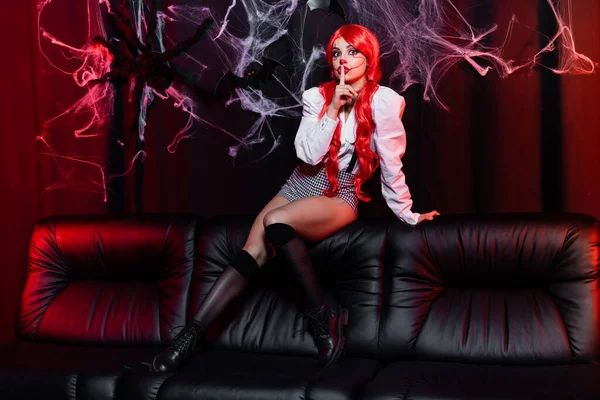 Mulher sexy no traje de palhaço halloween mostrando sinal de silêncio no sofá preto perto teia de aranha no fundo escuro — Fotografia de Stock
